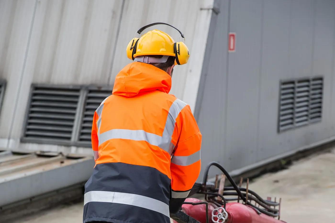 Arbeiter in orangefarbener Warnjacke mit Schutzhelm und Kapselgehörschutz zieht einen Gasflaschenwagen über einen Betriebshof