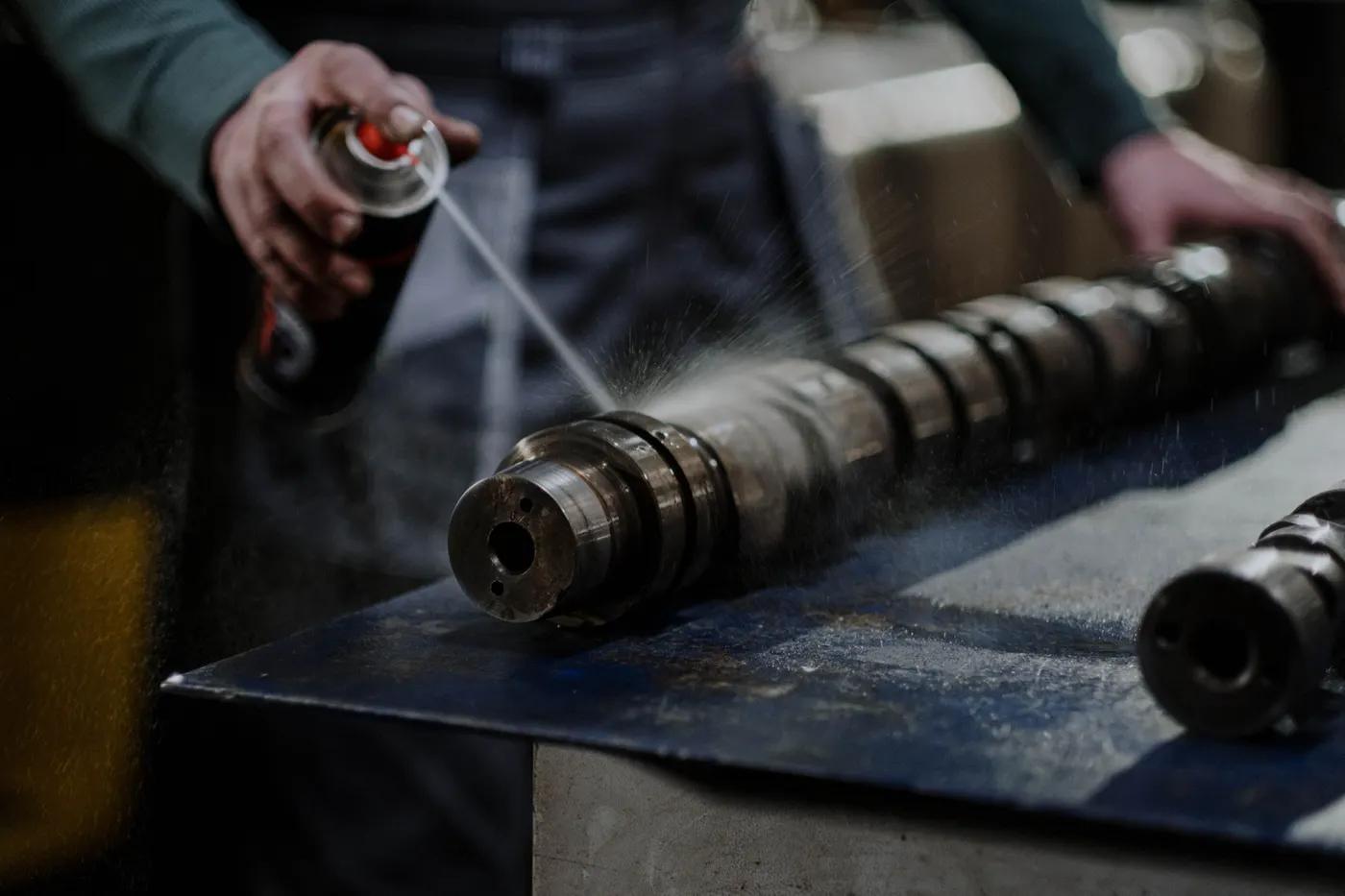 Hand sprüht ein technisches Spray aus der Dose auf eine Nockenwelle auf der Werkbank